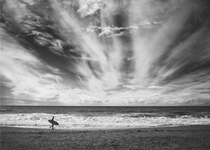A dramatic black and white photograph of a lone surfer silhouetted against the sea, walking along a sandy beach. The sky is filled with spectacular, radiating clouds that stretch across the horizon, creating a powerful and moody atmosphere. The image is enclosed in a silver frame. Poster