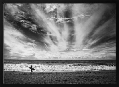 A dramatic black and white photograph of a lone surfer silhouetted against the sea, walking along a sandy beach. The sky is filled with spectacular, radiating clouds that stretch across the horizon, creating a powerful and moody atmosphere. The image is enclosed in a silver frame. Poster