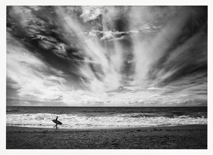 A dramatic black and white photograph of a lone surfer silhouetted against the sea, walking along a sandy beach. The sky is filled with spectacular, radiating clouds that stretch across the horizon, creating a powerful and moody atmosphere. The image is enclosed in a silver frame. Poster