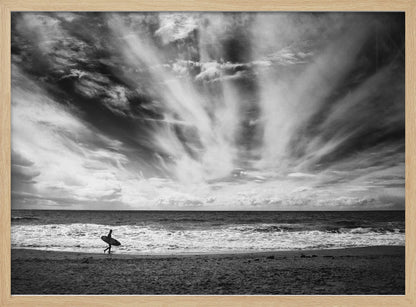 A dramatic black and white photograph of a lone surfer silhouetted against the sea, walking along a sandy beach. The sky is filled with spectacular, radiating clouds that stretch across the horizon, creating a powerful and moody atmosphere. The image is enclosed in a silver frame. Poster