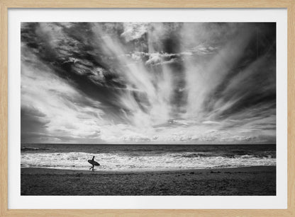 A dramatic black and white photograph of a lone surfer silhouetted against the sea, walking along a sandy beach. The sky is filled with spectacular, radiating clouds that stretch across the horizon, creating a powerful and moody atmosphere. The image is enclosed in a silver frame. Poster