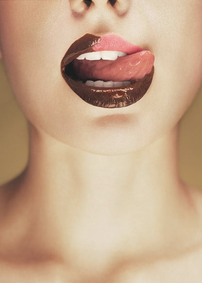A close-up photograph of a woman's mouth with two-toned lipstick. The lips are mostly covered in glossy dark brown, with the upper right section in light pink. Her tongue is out, licking the pink part of her lip, revealing her white teeth. Print