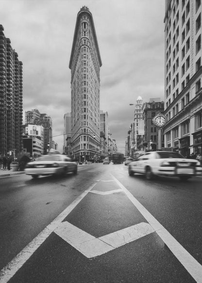 A dynamic black and white, low-angle photograph of the iconic Flatiron Building in New York City. The street in the foreground has bold white lines leading the eye to the building, while cars on either side are blurred with motion, capturing the city's fast pace under a cloudy sky. Decor