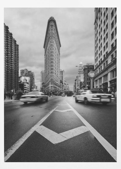 A dynamic black and white, low-angle photograph of the iconic Flatiron Building in New York City. The street in the foreground has bold white lines leading the eye to the building, while cars on either side are blurred with motion, capturing the city's fast pace under a cloudy sky. Decor