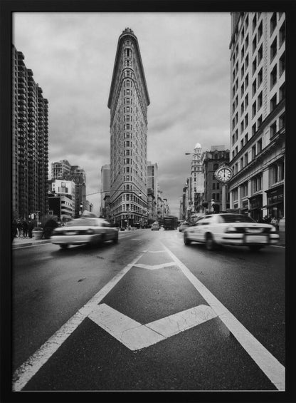 A dynamic black and white, low-angle photograph of the iconic Flatiron Building in New York City. The street in the foreground has bold white lines leading the eye to the building, while cars on either side are blurred with motion, capturing the city's fast pace under a cloudy sky. Decor