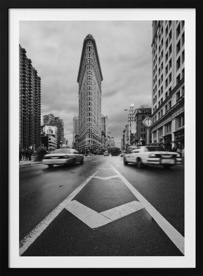 A dynamic black and white, low-angle photograph of the iconic Flatiron Building in New York City. The street in the foreground has bold white lines leading the eye to the building, while cars on either side are blurred with motion, capturing the city's fast pace under a cloudy sky. Decor