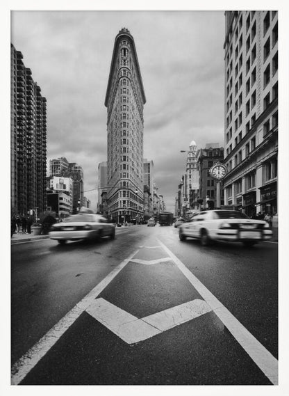 A dynamic black and white, low-angle photograph of the iconic Flatiron Building in New York City. The street in the foreground has bold white lines leading the eye to the building, while cars on either side are blurred with motion, capturing the city's fast pace under a cloudy sky. Decor