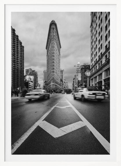 A dynamic black and white, low-angle photograph of the iconic Flatiron Building in New York City. The street in the foreground has bold white lines leading the eye to the building, while cars on either side are blurred with motion, capturing the city's fast pace under a cloudy sky. Decor
