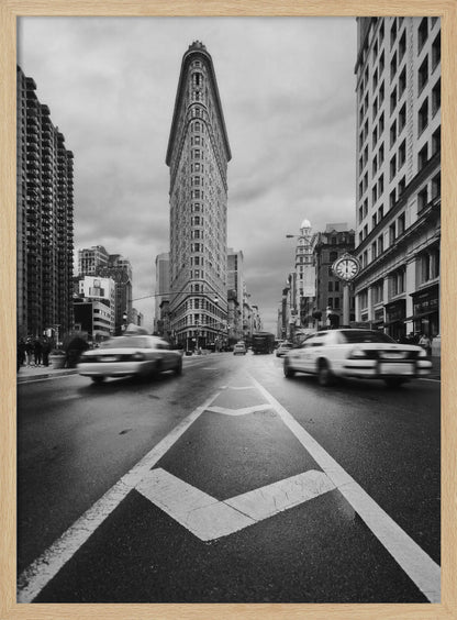 A dynamic black and white, low-angle photograph of the iconic Flatiron Building in New York City. The street in the foreground has bold white lines leading the eye to the building, while cars on either side are blurred with motion, capturing the city's fast pace under a cloudy sky. Decor
