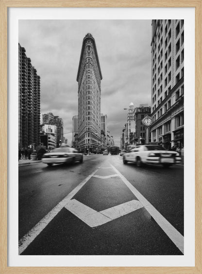 A dynamic black and white, low-angle photograph of the iconic Flatiron Building in New York City. The street in the foreground has bold white lines leading the eye to the building, while cars on either side are blurred with motion, capturing the city's fast pace under a cloudy sky. Decor