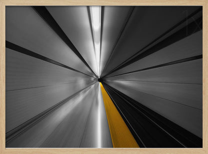 An abstract, long-exposure photograph from a first-person perspective inside a tunnel, creating a motion-blurred effect. The scene is mostly black and white, with lines radiating from a central vanishing point, but a single, bold yellow line runs down the center, adding a pop of selective color. The artwork is presented in a silver frame. Print