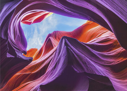 A low-angle photograph looking up from inside a vibrant slot canyon. The smooth, flowing sandstone walls are colored in rich shades of purple, red, and orange, curving around an opening that reveals a bright blue sky with wispy clouds. The image is presented within a silver-colored frame. Wall Art