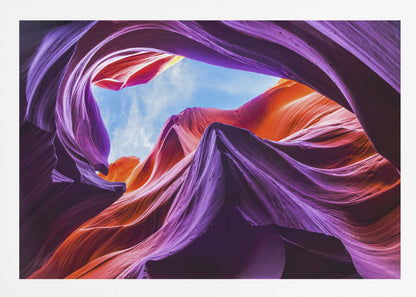 A low-angle photograph looking up from inside a vibrant slot canyon. The smooth, flowing sandstone walls are colored in rich shades of purple, red, and orange, curving around an opening that reveals a bright blue sky with wispy clouds. The image is presented within a silver-colored frame. Wall Art