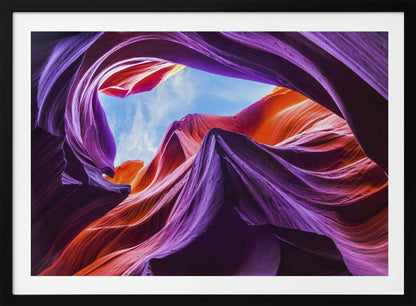A low-angle photograph looking up from inside a vibrant slot canyon. The smooth, flowing sandstone walls are colored in rich shades of purple, red, and orange, curving around an opening that reveals a bright blue sky with wispy clouds. The image is presented within a silver-colored frame. Wall Art