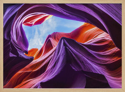 A low-angle photograph looking up from inside a vibrant slot canyon. The smooth, flowing sandstone walls are colored in rich shades of purple, red, and orange, curving around an opening that reveals a bright blue sky with wispy clouds. The image is presented within a silver-colored frame. Wall Art