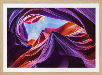 A low-angle photograph looking up from inside a vibrant slot canyon. The smooth, flowing sandstone walls are colored in rich shades of purple, red, and orange, curving around an opening that reveals a bright blue sky with wispy clouds. The image is presented within a silver-colored frame. Wall Art