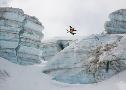 A framed photo of a skier in mid-air, jumping over a snowy crevasse between large, layered glacier formations under an overcast sky. The skier wears a black top and orange pants. Wall Art