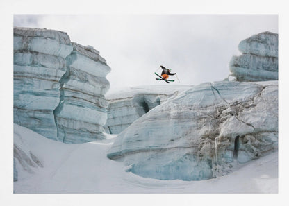 A framed photo of a skier in mid-air, jumping over a snowy crevasse between large, layered glacier formations under an overcast sky. The skier wears a black top and orange pants. Wall Art