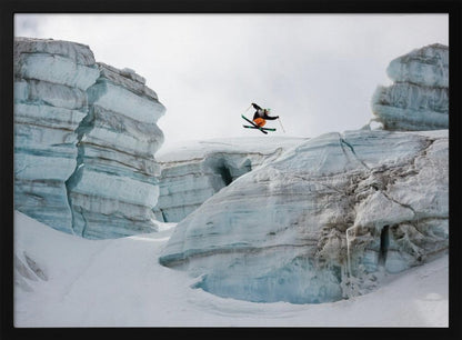A framed photo of a skier in mid-air, jumping over a snowy crevasse between large, layered glacier formations under an overcast sky. The skier wears a black top and orange pants. Wall Art