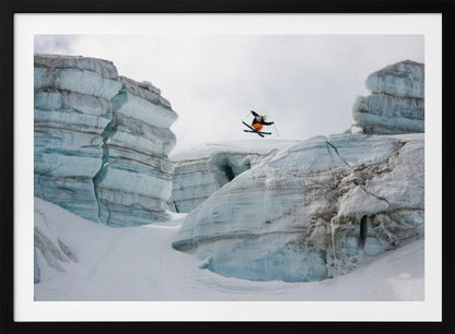 A framed photo of a skier in mid-air, jumping over a snowy crevasse between large, layered glacier formations under an overcast sky. The skier wears a black top and orange pants. Wall Art