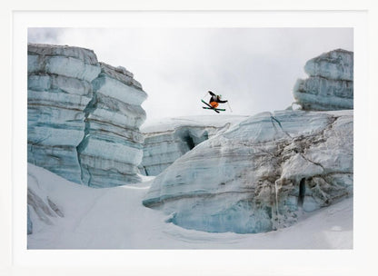 A framed photo of a skier in mid-air, jumping over a snowy crevasse between large, layered glacier formations under an overcast sky. The skier wears a black top and orange pants. Wall Art