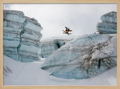A framed photo of a skier in mid-air, jumping over a snowy crevasse between large, layered glacier formations under an overcast sky. The skier wears a black top and orange pants. Wall Art