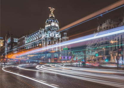 A dynamic long-exposure night photograph of the Metropolis Building in Madrid, with vibrant streaks of white, orange, and blue light from traffic swirling through the city streets. Artwork