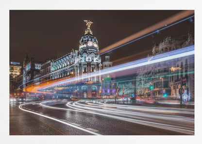 A dynamic long-exposure night photograph of the Metropolis Building in Madrid, with vibrant streaks of white, orange, and blue light from traffic swirling through the city streets. Artwork