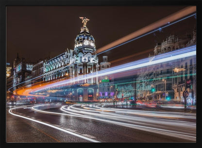 A dynamic long-exposure night photograph of the Metropolis Building in Madrid, with vibrant streaks of white, orange, and blue light from traffic swirling through the city streets. Artwork