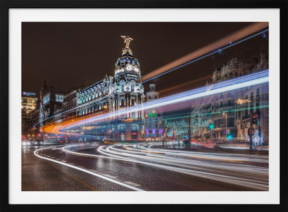 A dynamic long-exposure night photograph of the Metropolis Building in Madrid, with vibrant streaks of white, orange, and blue light from traffic swirling through the city streets. Artwork