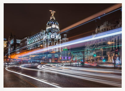 A dynamic long-exposure night photograph of the Metropolis Building in Madrid, with vibrant streaks of white, orange, and blue light from traffic swirling through the city streets. Artwork