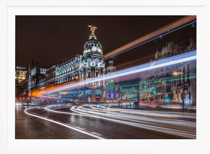 A dynamic long-exposure night photograph of the Metropolis Building in Madrid, with vibrant streaks of white, orange, and blue light from traffic swirling through the city streets. Artwork