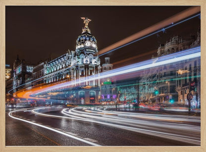 A dynamic long-exposure night photograph of the Metropolis Building in Madrid, with vibrant streaks of white, orange, and blue light from traffic swirling through the city streets. Artwork