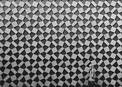 A black and white photograph of a lone person walking in front of a massive wall covered in a dizzying geometric pattern of tilted, textured gray quadrilaterals on a black background. The figure in the bottom right corner is dwarfed by the scale of the abstract architectural design. The image has a silver-gray frame. Artwork