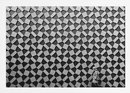 A black and white photograph of a lone person walking in front of a massive wall covered in a dizzying geometric pattern of tilted, textured gray quadrilaterals on a black background. The figure in the bottom right corner is dwarfed by the scale of the abstract architectural design. The image has a silver-gray frame. Artwork