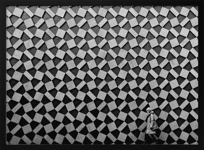 A black and white photograph of a lone person walking in front of a massive wall covered in a dizzying geometric pattern of tilted, textured gray quadrilaterals on a black background. The figure in the bottom right corner is dwarfed by the scale of the abstract architectural design. The image has a silver-gray frame. Artwork