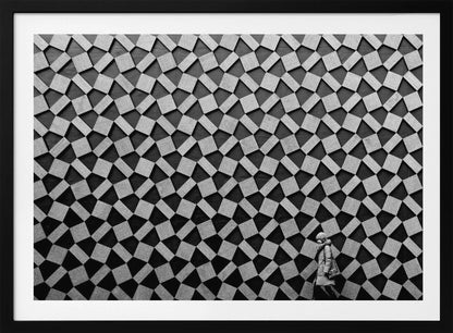 A black and white photograph of a lone person walking in front of a massive wall covered in a dizzying geometric pattern of tilted, textured gray quadrilaterals on a black background. The figure in the bottom right corner is dwarfed by the scale of the abstract architectural design. The image has a silver-gray frame. Artwork
