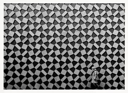 A black and white photograph of a lone person walking in front of a massive wall covered in a dizzying geometric pattern of tilted, textured gray quadrilaterals on a black background. The figure in the bottom right corner is dwarfed by the scale of the abstract architectural design. The image has a silver-gray frame. Artwork