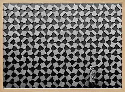 A black and white photograph of a lone person walking in front of a massive wall covered in a dizzying geometric pattern of tilted, textured gray quadrilaterals on a black background. The figure in the bottom right corner is dwarfed by the scale of the abstract architectural design. The image has a silver-gray frame. Artwork