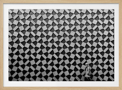 A black and white photograph of a lone person walking in front of a massive wall covered in a dizzying geometric pattern of tilted, textured gray quadrilaterals on a black background. The figure in the bottom right corner is dwarfed by the scale of the abstract architectural design. The image has a silver-gray frame. Artwork