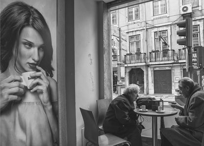 A framed, black and white photograph capturing a scene of juxtaposition. On the left, a large poster shows a glamorous young woman holding a coffee cup. On the right, seen through a cafe window, an elderly couple sits at a small table, also with coffee cups, engaged in conversation against a backdrop of an old European city street. Poster