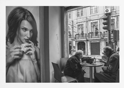 A framed, black and white photograph capturing a scene of juxtaposition. On the left, a large poster shows a glamorous young woman holding a coffee cup. On the right, seen through a cafe window, an elderly couple sits at a small table, also with coffee cups, engaged in conversation against a backdrop of an old European city street. Poster