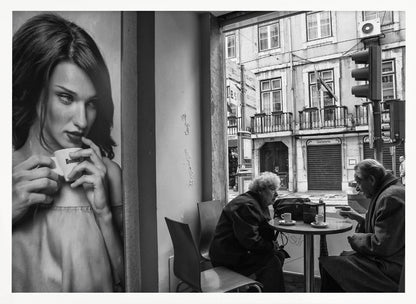 A framed, black and white photograph capturing a scene of juxtaposition. On the left, a large poster shows a glamorous young woman holding a coffee cup. On the right, seen through a cafe window, an elderly couple sits at a small table, also with coffee cups, engaged in conversation against a backdrop of an old European city street. Poster