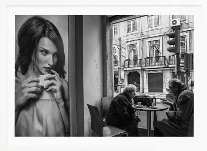 A framed, black and white photograph capturing a scene of juxtaposition. On the left, a large poster shows a glamorous young woman holding a coffee cup. On the right, seen through a cafe window, an elderly couple sits at a small table, also with coffee cups, engaged in conversation against a backdrop of an old European city street. Poster