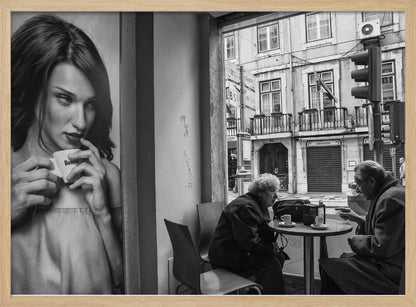 A framed, black and white photograph capturing a scene of juxtaposition. On the left, a large poster shows a glamorous young woman holding a coffee cup. On the right, seen through a cafe window, an elderly couple sits at a small table, also with coffee cups, engaged in conversation against a backdrop of an old European city street. Poster