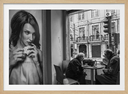 A framed, black and white photograph capturing a scene of juxtaposition. On the left, a large poster shows a glamorous young woman holding a coffee cup. On the right, seen through a cafe window, an elderly couple sits at a small table, also with coffee cups, engaged in conversation against a backdrop of an old European city street. Poster