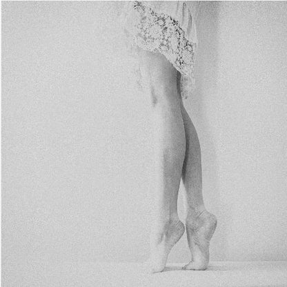 A framed, grainy black and white photograph of a dancer's legs and feet. The dancer stands on tiptoes against a white wall, wearing a delicate lace skirt. Poster