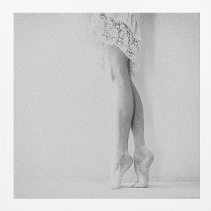 A framed, grainy black and white photograph of a dancer's legs and feet. The dancer stands on tiptoes against a white wall, wearing a delicate lace skirt. Poster