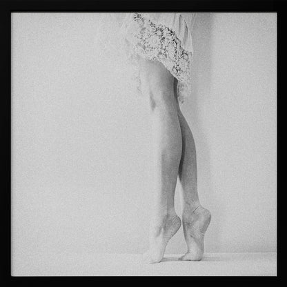 A framed, grainy black and white photograph of a dancer's legs and feet. The dancer stands on tiptoes against a white wall, wearing a delicate lace skirt. Poster