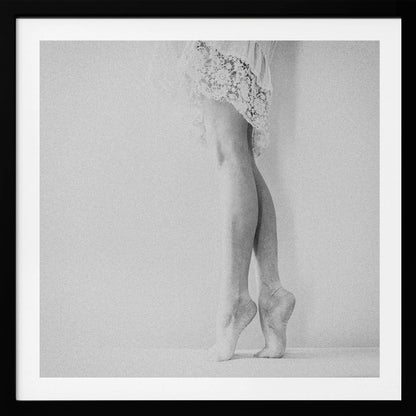 A framed, grainy black and white photograph of a dancer's legs and feet. The dancer stands on tiptoes against a white wall, wearing a delicate lace skirt. Poster